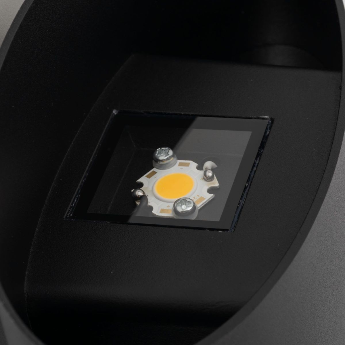 Kinkiet Verto 1xLED 22W IP54 w kolorze czarnym, widoczny w zbliżeniu na źródło światła LED. Elementy montażowe są widoczne, a lampa ma prosty, nowoczesny kształt. Ujęcie z góry, na tle ciemnego materiału.
