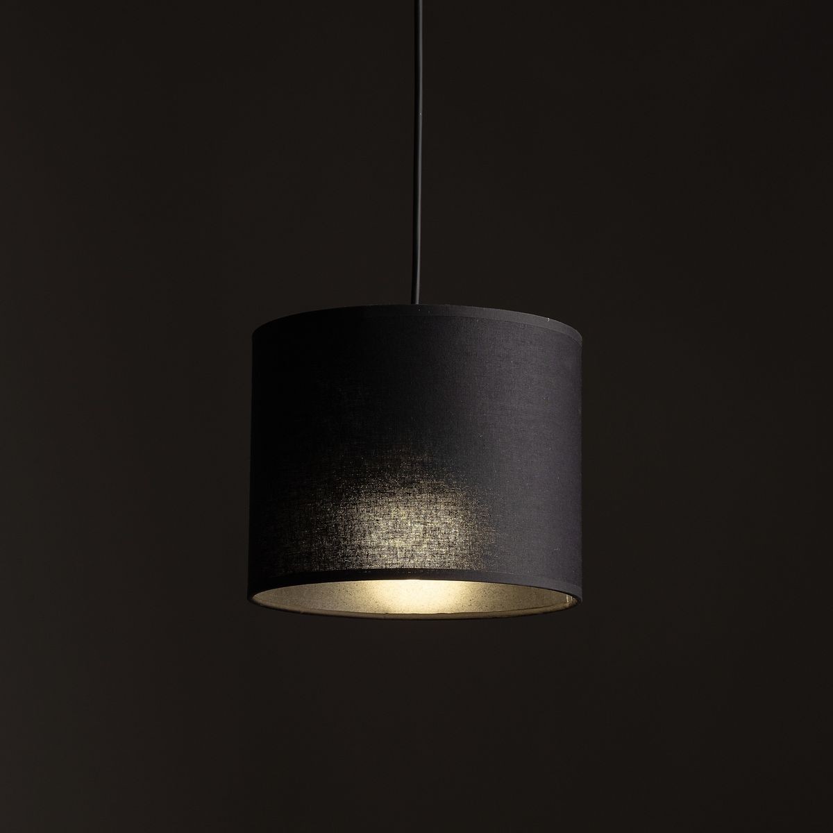 Lampa wisząca Tracer D20 w czarnym kolorze z materiałowym kloszem w kształcie cylindra, zawieszona na cienkim przewodzie. Wewnątrz klosza znajduje się żarówka E27, emitująca przyjemne światło. Tło ciemne, podkreślające elegancję lampy.