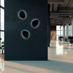 Kinkiet Stone 3xG9 w kolorze czarnym, montowany na ścianie, z trzema otworami w kształcie kropli. Oświetlenie emituje subtelne światło wokół otworów, w nowoczesnym biurowym wnętrzu z dużymi oknami i roślinnością.