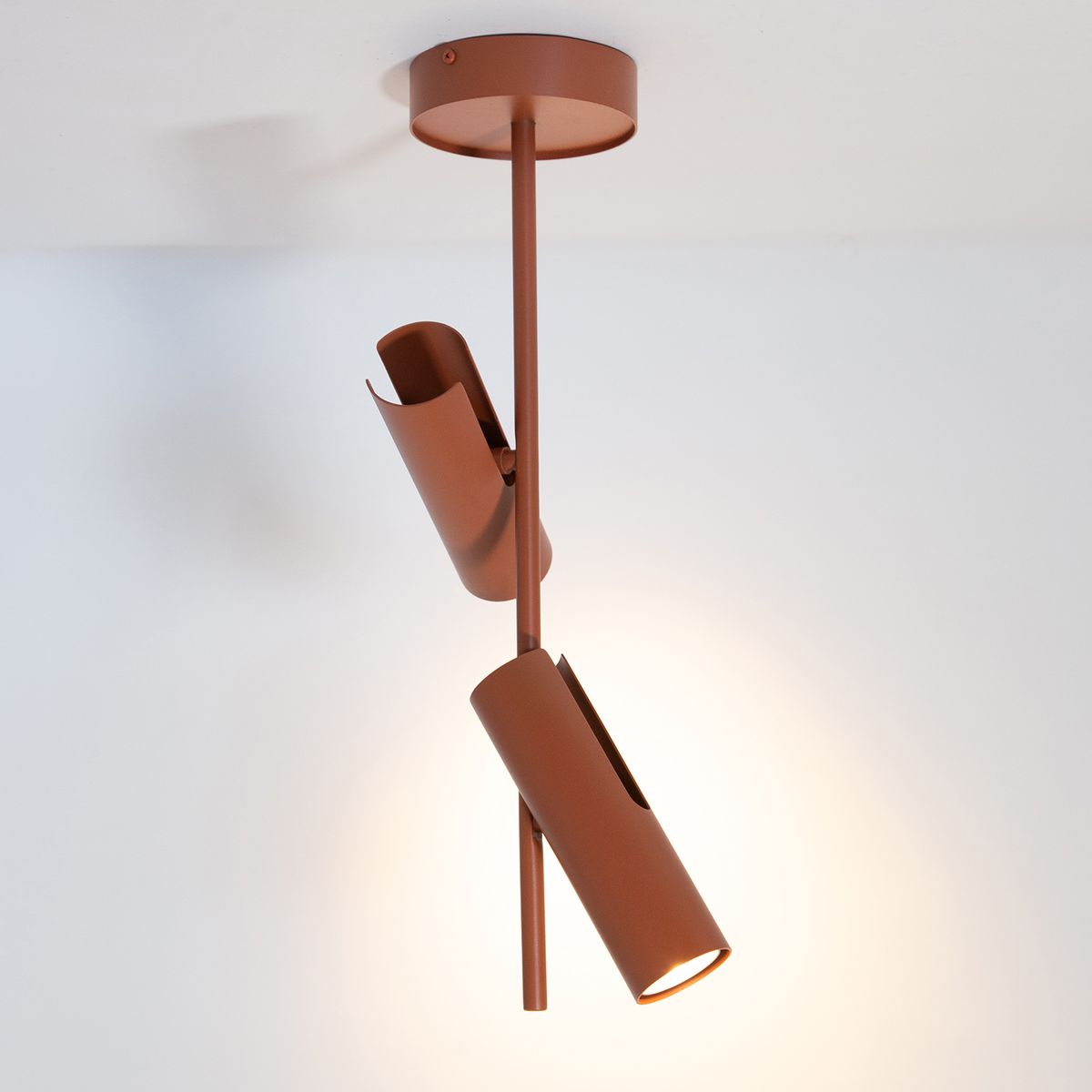 Lampa wisząca Leda, ceglana, z dwoma regulowanymi reflektorami GU10. Stojąca na metalowym stelażu, z widocznym okrągłym mocowaniem sufitowym. Reflektory w kształcie cylindrów, z otwartymi końcami, emitują ciepłe światło, tworząc nowoczesny akcent.
