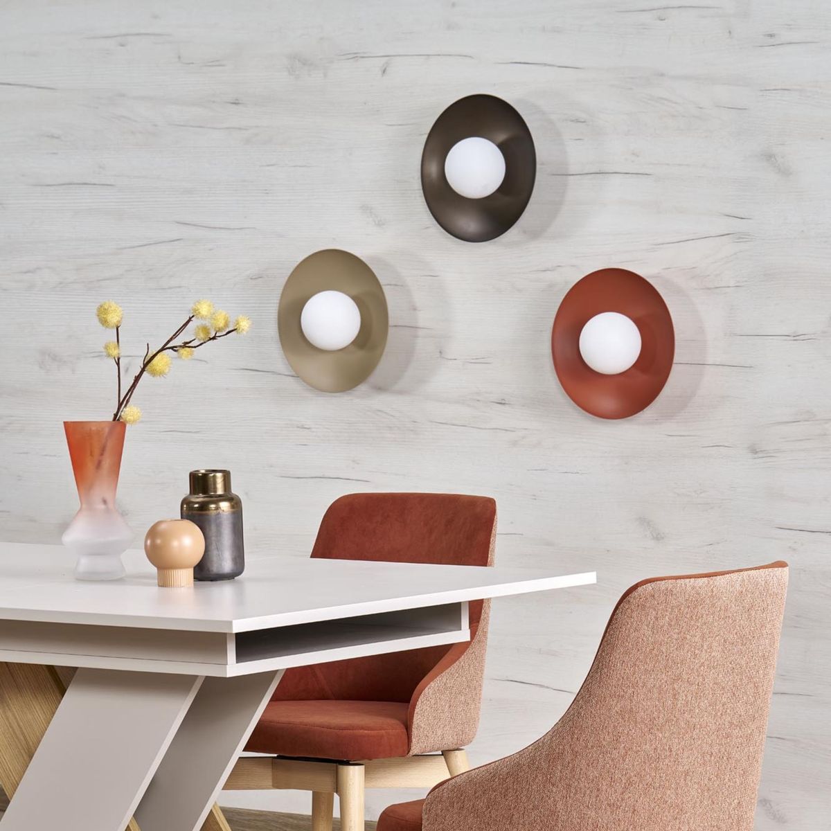 Kinkiet Slate w beżowym kolorze, z okrągłą, matową oprawą oraz białą kulistą żarówką, zamontowany na ścianie. W tle widoczny stół i krzesła w nowoczesnym stylu, na tle jasnej, drewnopodobnej ściany.