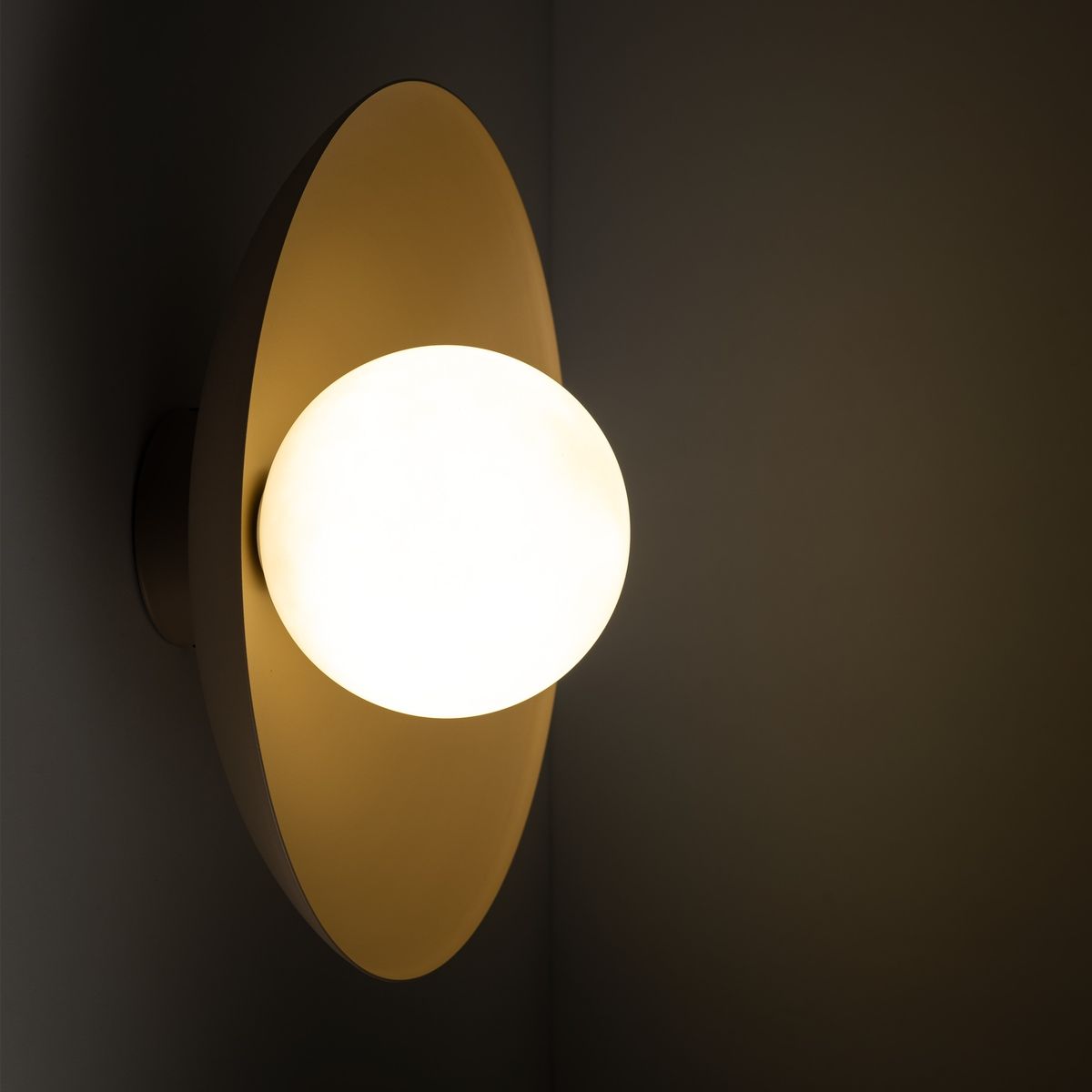 Beżowy kinkiet Slate z okrągłą, matową żarówką o ciepłym świetle, zamontowany na ścianie. Kinkiet ma elegancki, minimalistyczny kształt z dużą, owalną podstawą, tworząc nowoczesny akcent w aranżacji wnętrza.