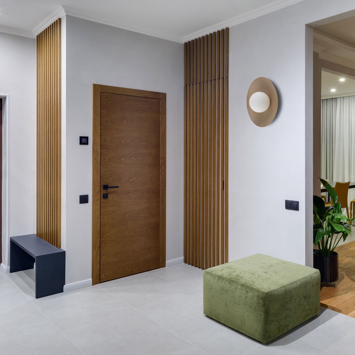 Kinkiet Slate w kolorze beżowym, o okrągłym kształcie, zamontowany na ścianie. W tle widoczna drewniana drzwi oraz zielony puf. Oświetlenie w neutralnym wnętrzu z jasnymi ścianami i podłogą.