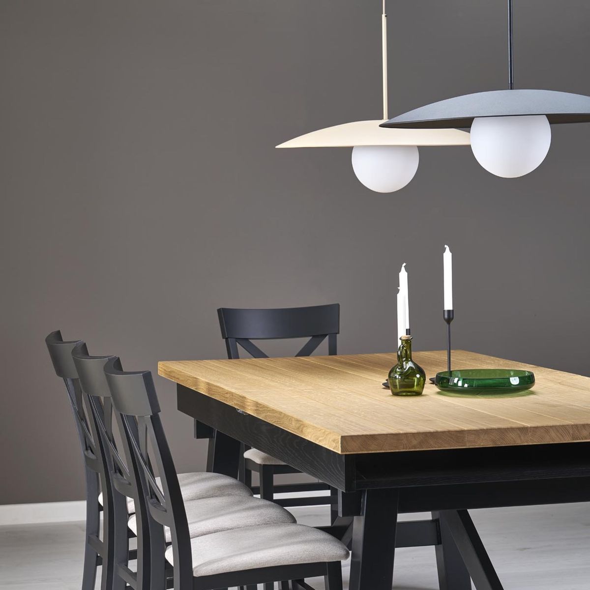 Beżowa lampa wisząca Slate z jednym źródłem światła E27, zawieszona nad drewnianym stołem, w otoczeniu czterech czarnych krzeseł. W tle szare ściany, na stole zielona butelka i świece, tworzące przytulną aranżację.