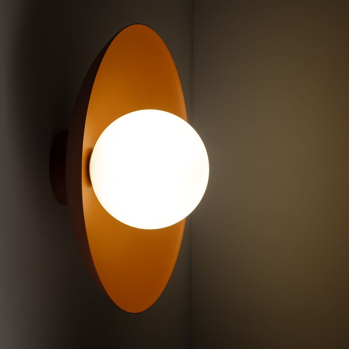 Kinkiet Slate 1xG9 w ceglastym kolorze, z okrągłym, matowym kloszem w białym kolorze, zamontowany na ścianie. Wykonany z metalu, o nowoczesnym kształcie, z widocznym połączeniem z podstawą.