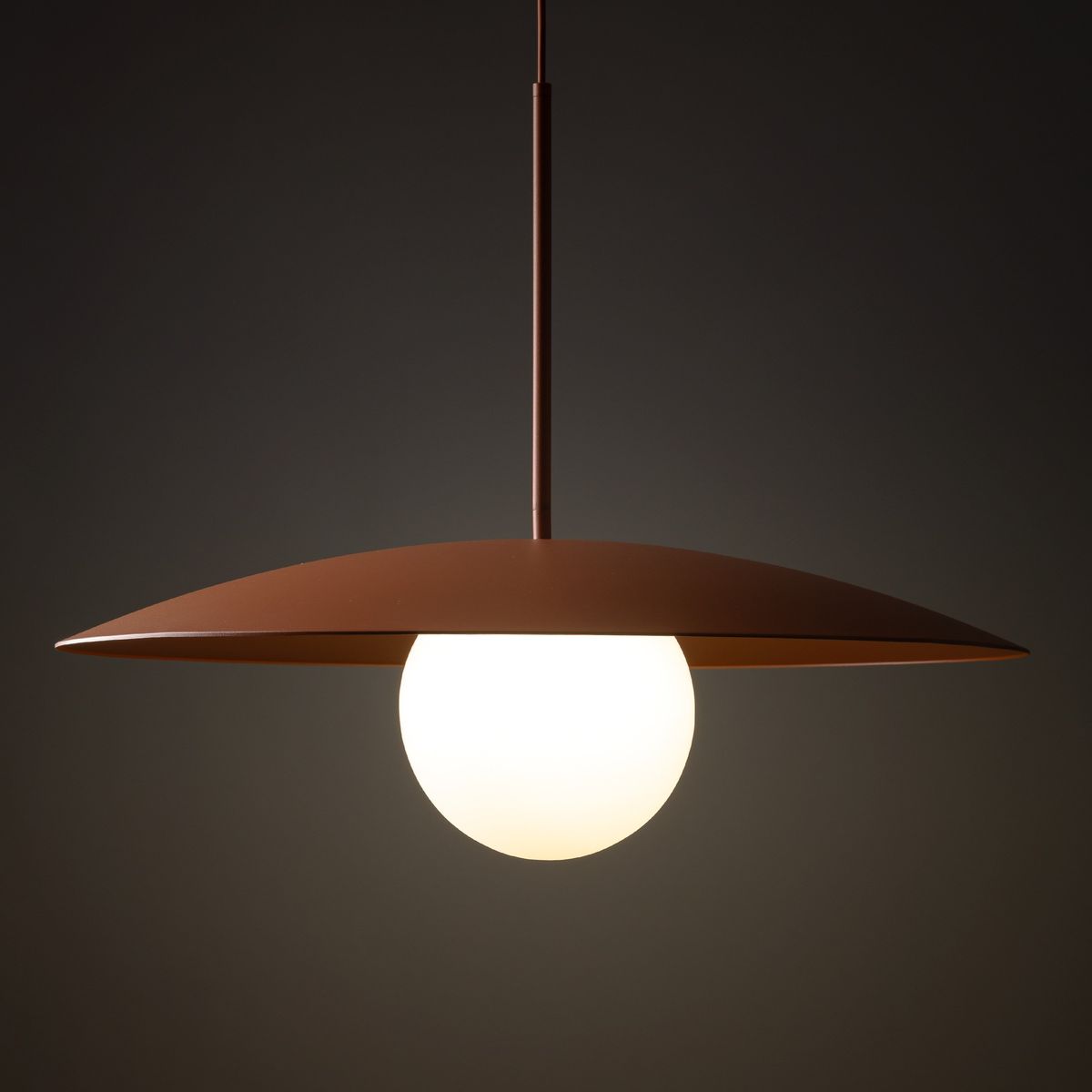 Lampa wisząca Slate w ceglastym kolorze z metalowym kloszem w kształcie talerza, zawieszona na cienkim przewodzie. W centrum znajduje się okrągła, matowa kula jako źródło światła. Tło ciemne, podkreślające elegancki design.