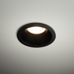 Czarna oprawa sufitowa do zabudowy Opti 1xGU10 IP64, zamontowana na białym suficie. Widoczny okrągły kształt z matowym wykończeniem, emitująca ciepłe światło. Idealna do oświetlenia wnętrz.