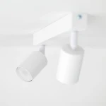 Lampa sufitowa Nex w kolorze białym z dwoma punktami świetlnymi typu GU10. Ujęcie z boku, pokazujące cylindryczne klosze i prostokątną podstawę. Elementy zamontowane pod sufitem, minimalistyczny design.