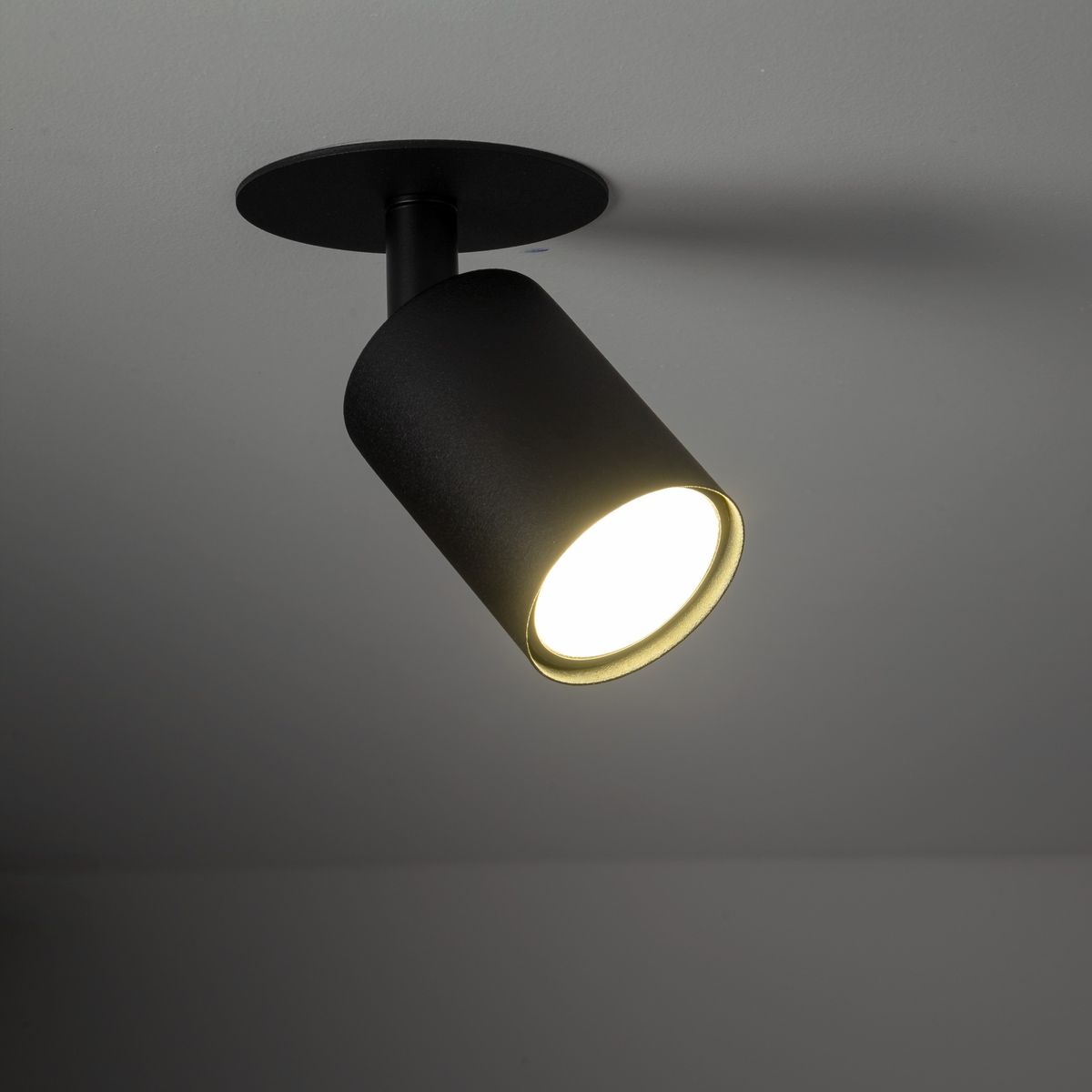 Lampa sufitowa podtynkowa Nex w kolorze czarnym, z pojedynczym źródłem światła GU10. Cylindryczny kształt, zamontowana na okrągłej podstawie. Oświetlenie skierowane w dół, na jednolitym tle.