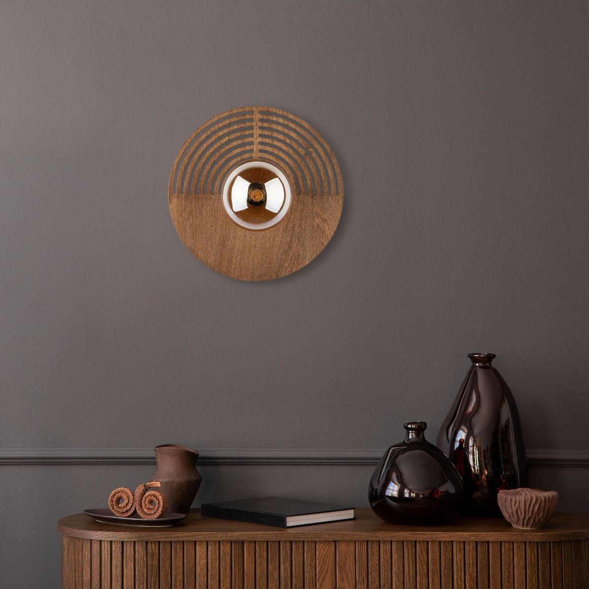 Kinkiet Lunar Wood D30 w kolorze drewna z okrągłą podstawą z wypukłym elementem w centralnej części. Ułożony na szarej ścianie, na tle drewnianego stolika z dekoracyjnymi naczyniami i książkami.