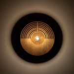 Kinkiet Lunar Wood D25 z drewna, z centralnym źródłem światła G9. Okrągła, brązowa obudowa z wycięciami w formie fal. Lampa zamontowana na ścianie, emitująca delikatne światło, tworząca ciekawe cienie na tle.