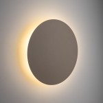 Kinkiet Luna 4xG9 w kształcie okręgu, o matowej, szarej powierzchni. Emisja światła tworzy delikatny efekt podświetlenia w odcieniach żółtego. Zamontowany na ścianie, widok frontalny.