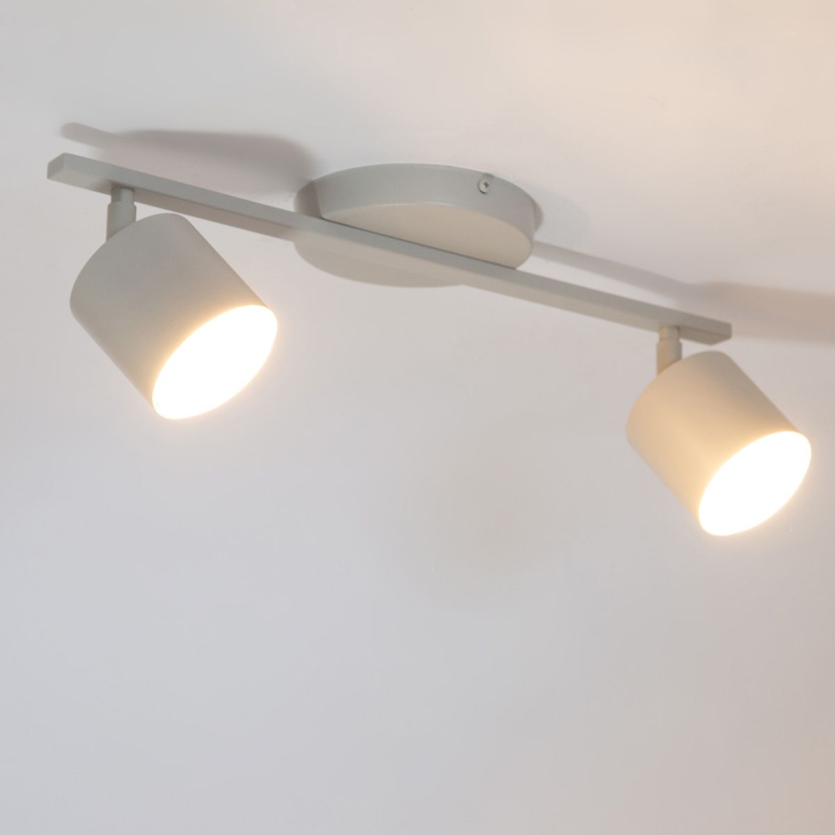Lampa sufitowa Mila z dwoma reflektorami GX53 w kolorze szarym beżowym. Zamontowana na prostokątnej podstawie, ukierunkowane klosze emitują światło, podkreślając minimalistyczny design. Ujęcie z dołu, w zbliżeniu na szczegóły.