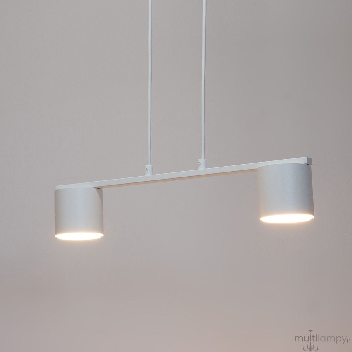 Lampa wisząca Mila 2xGX53 w kolorze białym, składająca się z dwóch cylindrycznych kloszy zamocowanych na poziomej ramie. Klosze emitują światło, a lampa zawieszona jest na dwóch białych przewodach. Prosta, nowoczesna forma.