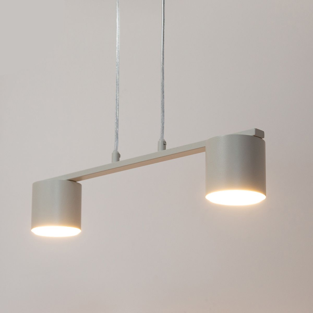 Lampa wisząca Mila w kolorze szarym beżowym, z dwoma cylindrycznymi kloszami na końcach prostokątnego ramienia. Zawieszona na dwóch cienkich linkach, emitująca jasne światło.