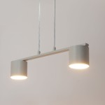 Lampa wisząca Mila w kolorze szarym beżowym, z dwoma cylindrycznymi kloszami na końcach prostokątnego ramienia. Zawieszona na dwóch cienkich linkach, emitująca jasne światło.