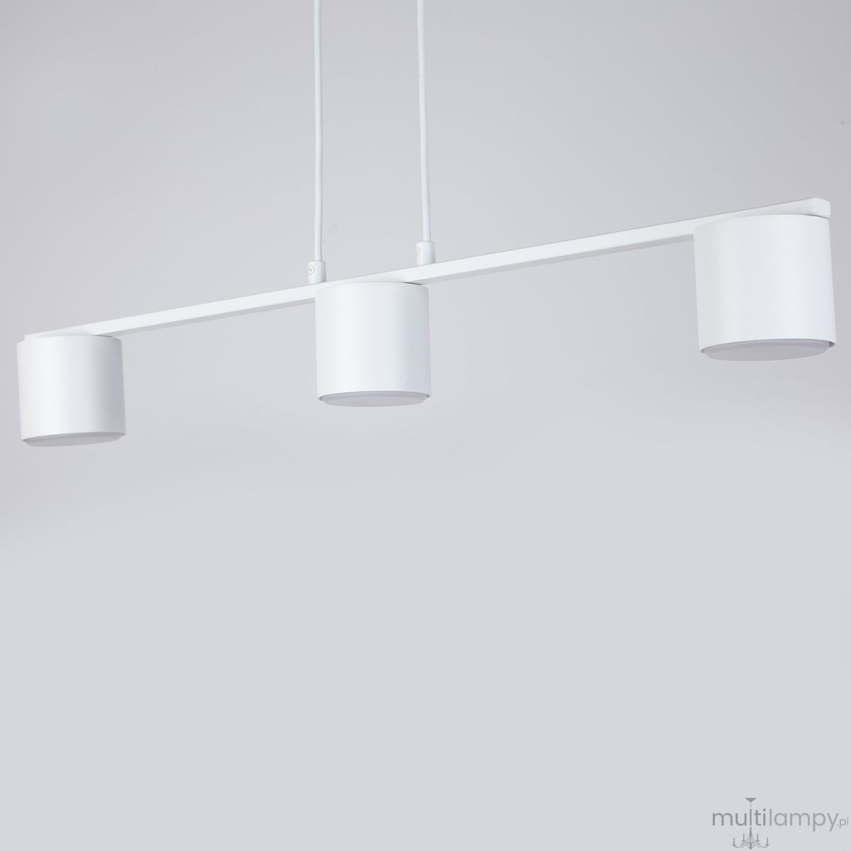 Lampa wisząca Mila w kolorze białym, z trzema cylindrycznymi kloszami na żarówki GX53. Produkt zawieszony na dwóch linkach, z minimalistycznym designem, idealny do nowoczesnych wnętrz. Tło w jednolitym, jasnym kolorze.