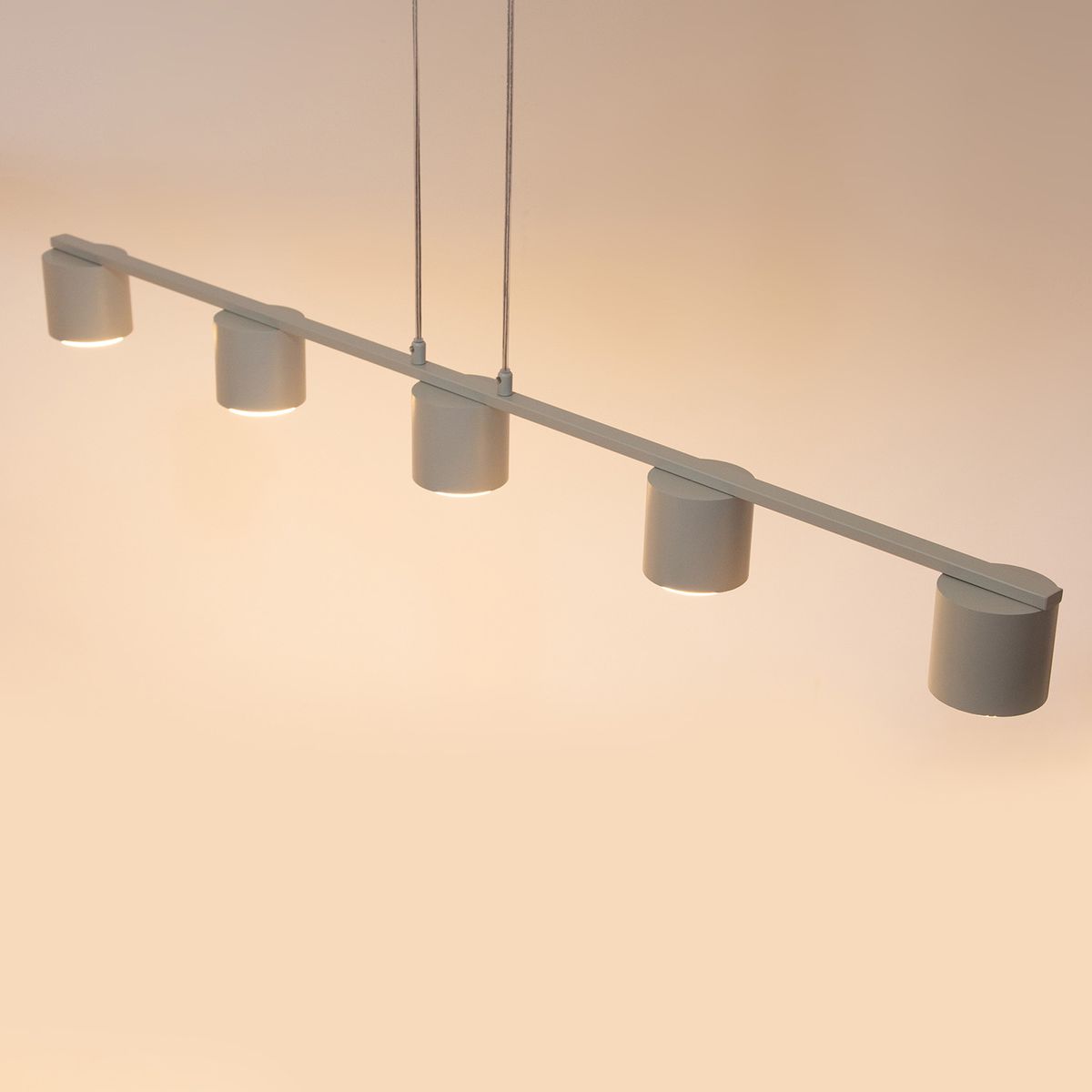 Lampa wisząca Mila w kolorze szaro-beżowym z pięcioma cylindrycznymi kloszami, zawieszona na prostym, metalowym stelażu. Klosze skierowane w dół, zawieszenie na dwóch sznurach. Na tle jasnym, minimalistyczny design.
