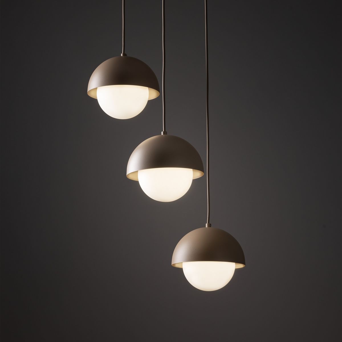 Lampa wisząca Bono z trzema kulistymi kloszami w kolorze beżowym, z białymi, szklanymi lampkami. Klosze mają półkolisty kształt i są zawieszone na cienkich przewodach, tworząc nowoczesny efekt wizualny na ciemnym tle.