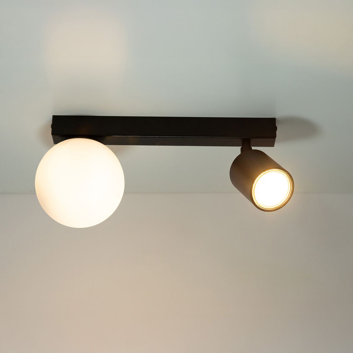 Czarna lampa sufitowa Tavira z dwoma źródłami światła: okrągła kula z białego szkła oraz reflektor w formie cylindra. Ujęcie z góry, lampa zamontowana na suficie, prezentuje nowoczesny design.