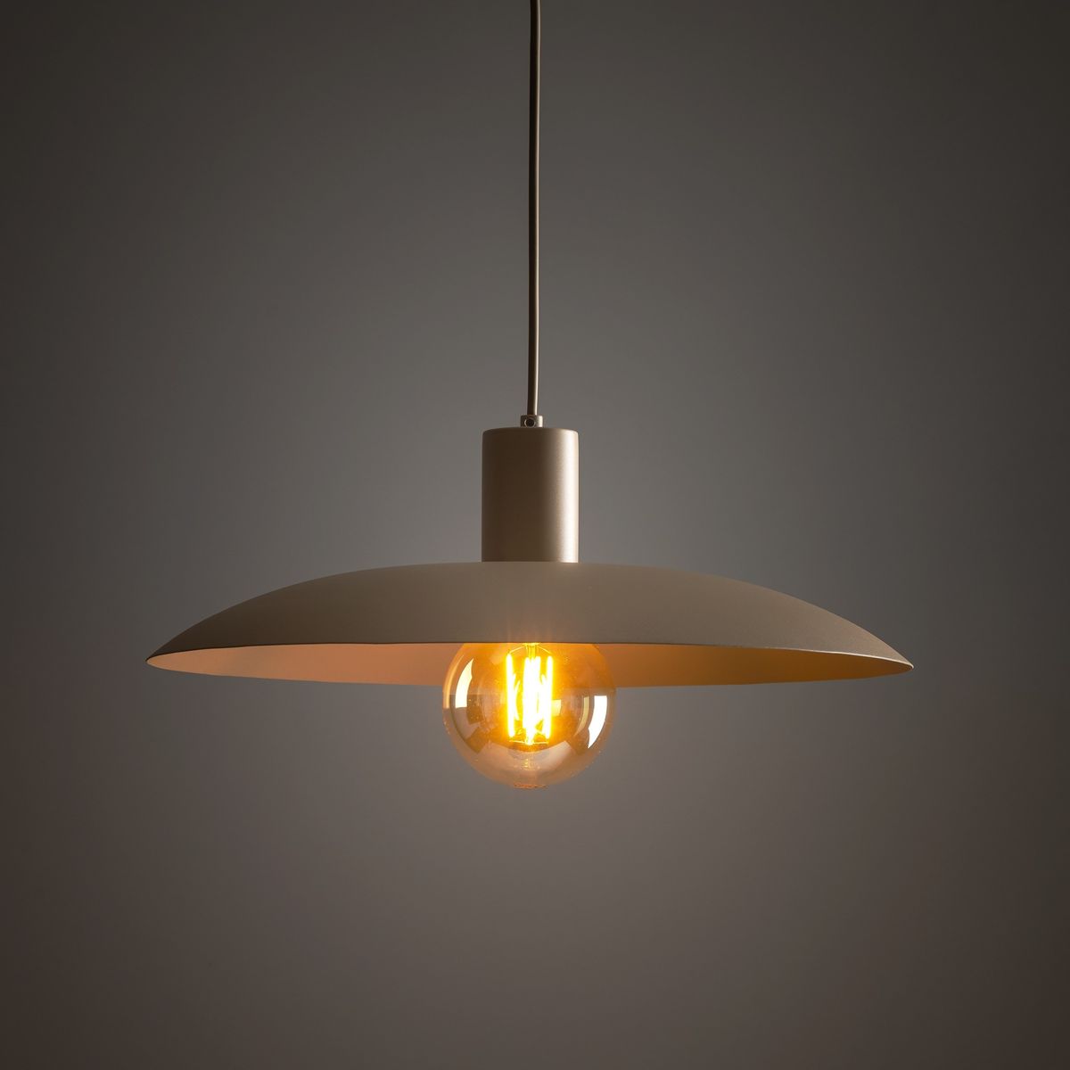 Lampa wisząca Astra z oprawą w kolorze szarym, z dużym, okrągłym kloszem i żarówką E27. Zawieszona na cienkim, metalowym przewodzie, emitująca ciepłe światło, idealna do nowoczesnych wnętrz. Tło w ciemnym odcieniu.