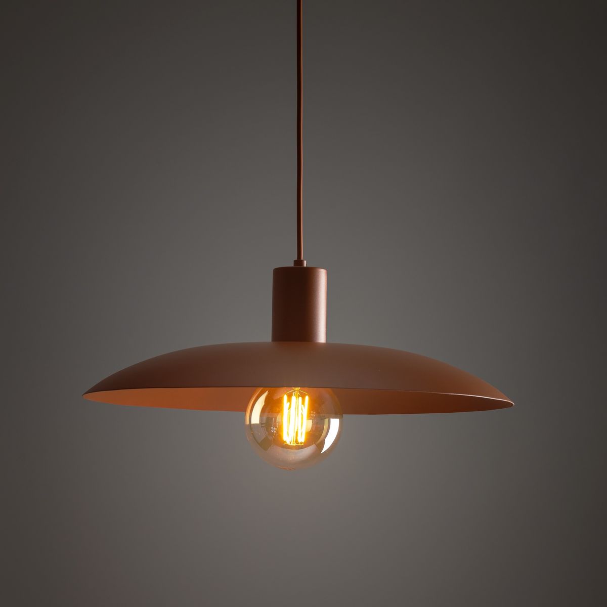 Lampa wisząca Astra w kolorze ceglanym z metalowym kloszem w kształcie talerza. Wyposażona w żarówkę E27, zawieszona na długim przewodzie. Tło ciemnoszare, podkreślające elegancki design.