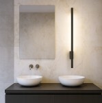 Linera kinkiet elewacyjny LED 9W 4000K w kolorze czarnym, zamontowany na ścianie. Produkt ma prostokątny kształt, emitując ciepłe światło. W tle widoczna umywalka i lustro, całość w aranżacji łazienkowej.