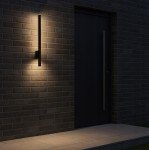 Kinkiet elewacyjny LED 9W w kolorze czarnym, umieszczony na ścianie z cegły. Prosty, smukły kształt z ciepłym białym światłem 4000K, zamontowany obok czarnych drzwi. Wysoka klasa ochrony IP44, odpowiedni do użytku na zewnątrz.