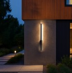 Linera kinkiet elewacyjny LED 9W 4000K w kolorze czarnym, zamontowany na szarej ścianie budynku, emituje ciepłe światło. Prosty, pionowy kształt z matowym wykończeniem, otoczony zielenią i ścieżką z płyt. Idealny do oświetlenia zewnętrznego.