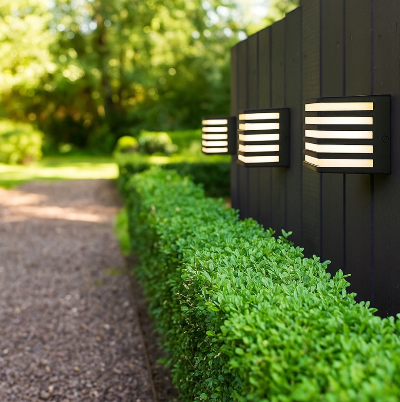 Kinkiet elewacyjny Cube II w kolorze czarnym, umieszczony na ścianie zewnętrznej, z białymi szczelinami, emitującym światło. W tle widoczna zielona żywopłot i ścieżka. Oświetlenie w nowoczesnym stylu, idealne do aranżacji ogrodowej.