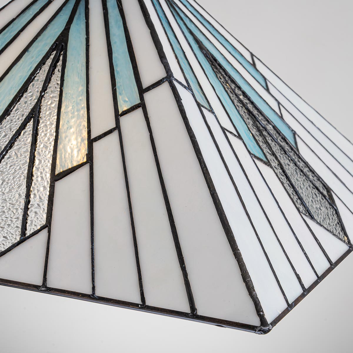 Lampa wisząca Alderley z kloszem w odcieniach bieli, błękitu i czerni, wykonana z witrażowego szkła. Zbliżenie na geometryczny wzór oraz antyczną brązową oprawę. Klosz ma kształt stożka, montowana na jednym źródle światła E27.