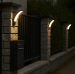 Kinkiet elewacyjny LED Grudo 12W 4000K w kolorze czarnym, zamontowany na kamiennej kolumnie ogrodzenia. Oświetlenie ma lekko zakrzywiony kształt, emitujące ciepłe światło, z widocznym czarnym pudełkiem na listy obok.