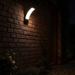 Kinkiet elewacyjny LED Grudo 12W 4000K w kolorze czarnym, zamontowany na ceglanej ścianie. Oświetlenie emituje ciepłe światło przez zaokrągloną, matową osłonę. W tle widoczne rośliny i drewniane drzwi. Klasa szczelności IP54.