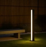 Lampa ogrodowa słupek Roma H60, czarna, o prostokątnym kształcie, z dwoma źródłami LED 24W 4000K. Ustawiona na trawie obok betonowej ławki, emituje ciepłe światło, tworząc przyjemną atmosferę w ogrodzie.