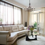 Lampa wisząca Windermere 6x E14 w kolorze złoto-brązowym, z sześcioma kloszami w kształcie świec. Zawieszona w jasnym wnętrzu z kanapą i szklanym stolikiem, otoczona zasłonami i roślinami.