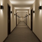 Czarny kinkiet elewacyjny Gantio z podwójnym źródłem LED 6W, 4000K, IP44, zamontowany na ścianie w wąskim korytarzu. Oświetlenie skierowane w dół, tworzące przyjemną atmosferę. W tle widoczna równa linia drzwi.