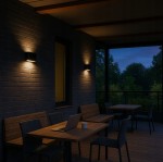 Kinkiet elewacyjny Gantio w kolorze czarnym, z dwoma źródłami LED 6W, 4000K. Montowany na ścianie, emituje ciepłe światło, oświetlając otoczenie. Widoczny w aranżacji z drewnianymi meblami na tarasie, w nocy. Ochrona IP44.