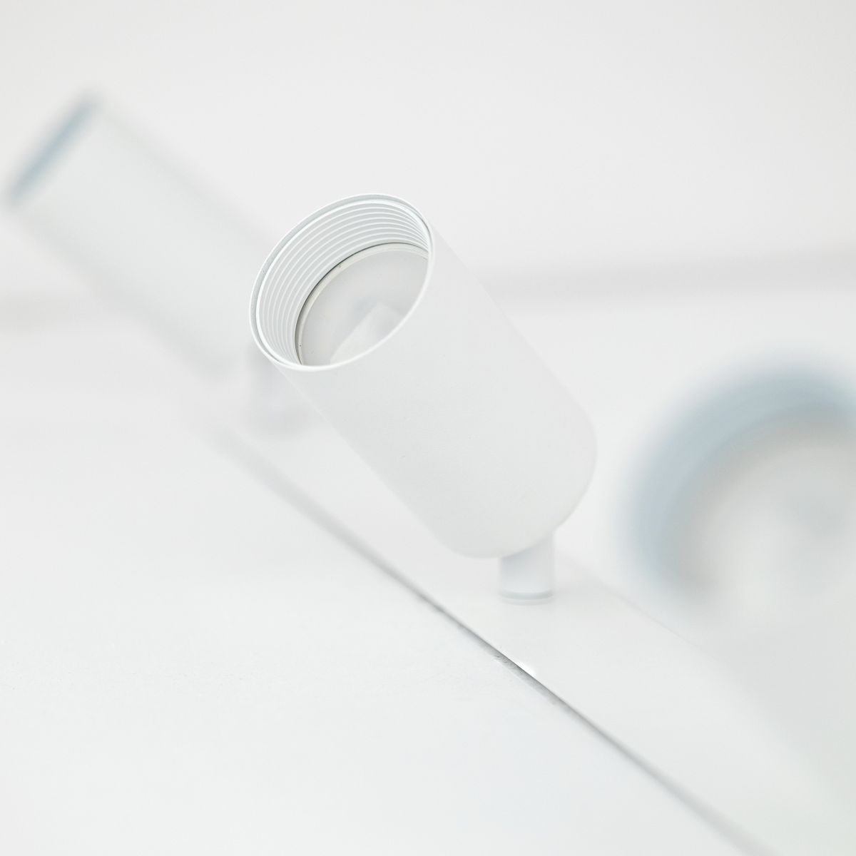 Lampa sufitowa podtynkowa Amis w białym kolorze z trzema oprawami na żarówki GU10. Ujęcie zbliżone na jeden z reflektorów, pokazujące jego cylindryczny kształt i gładką, matową powierzchnię. Na tle jasnej powierzchni, minimalistyczny design.