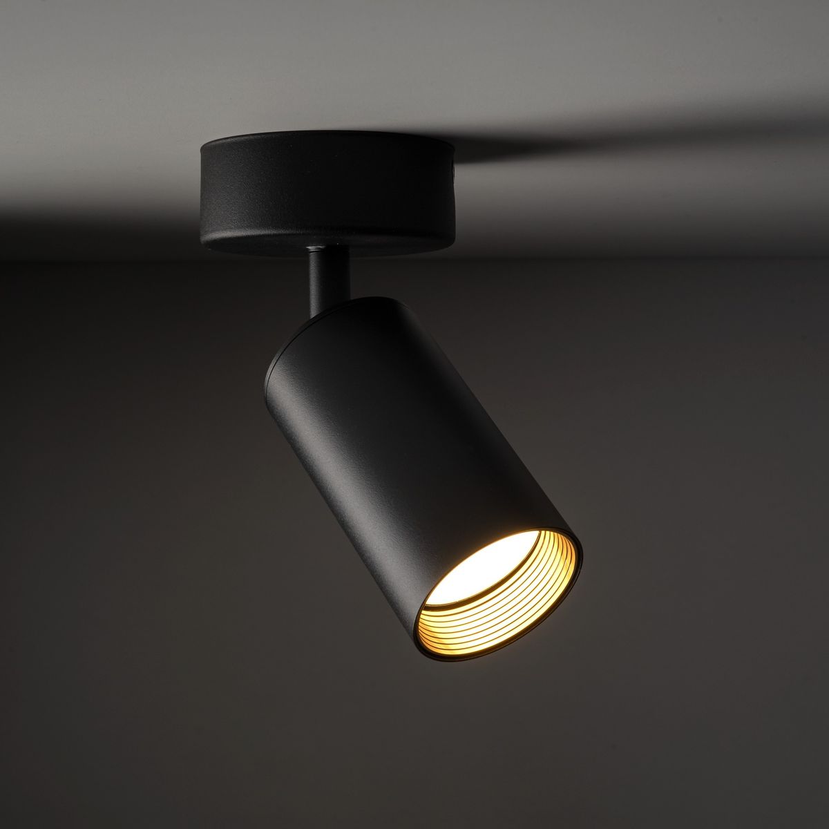 Lampa sufitowa Amis w kolorze czarnym z jednym gniazdem GU10. Ujęcie z boku, pokazujące cylindryczny klosz z odsłoniętym żarnikiem. Elementy metalowe, matowe wykończenie, montowana do sufitu.