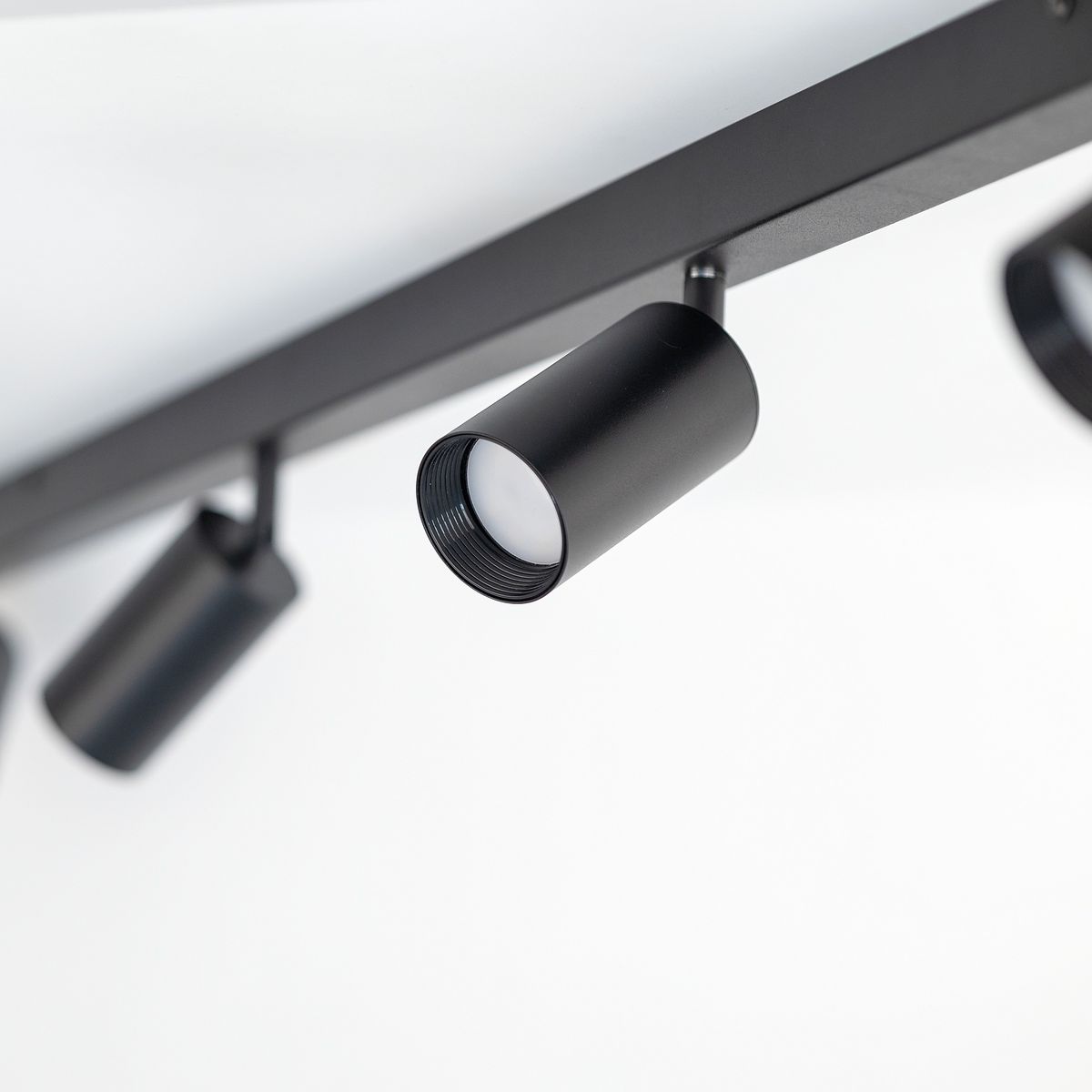 Lampa sufitowa Amis w kolorze czarnym, z czterema spotami na żarówki GU10. Ujęcie z boku, pokazujące jeden z reflektorów w matowym wykończeniu, zawieszony na prostokątnej podstawie. Tło białe, minimalistyczne.