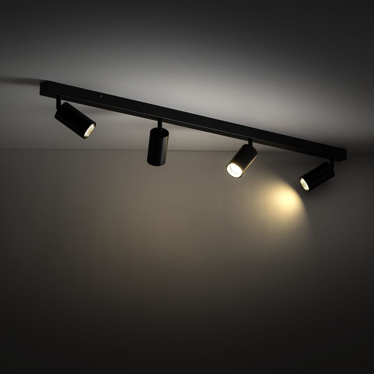 Czarna lampa sufitowa Amis z czterema reflektorami GU10, zamontowana na prostokątnej listwie. Reflektory skierowane w różne strony, na gładkim tle. Widać różne natężenie światła, podkreślające funkcjonalność oświetlenia.