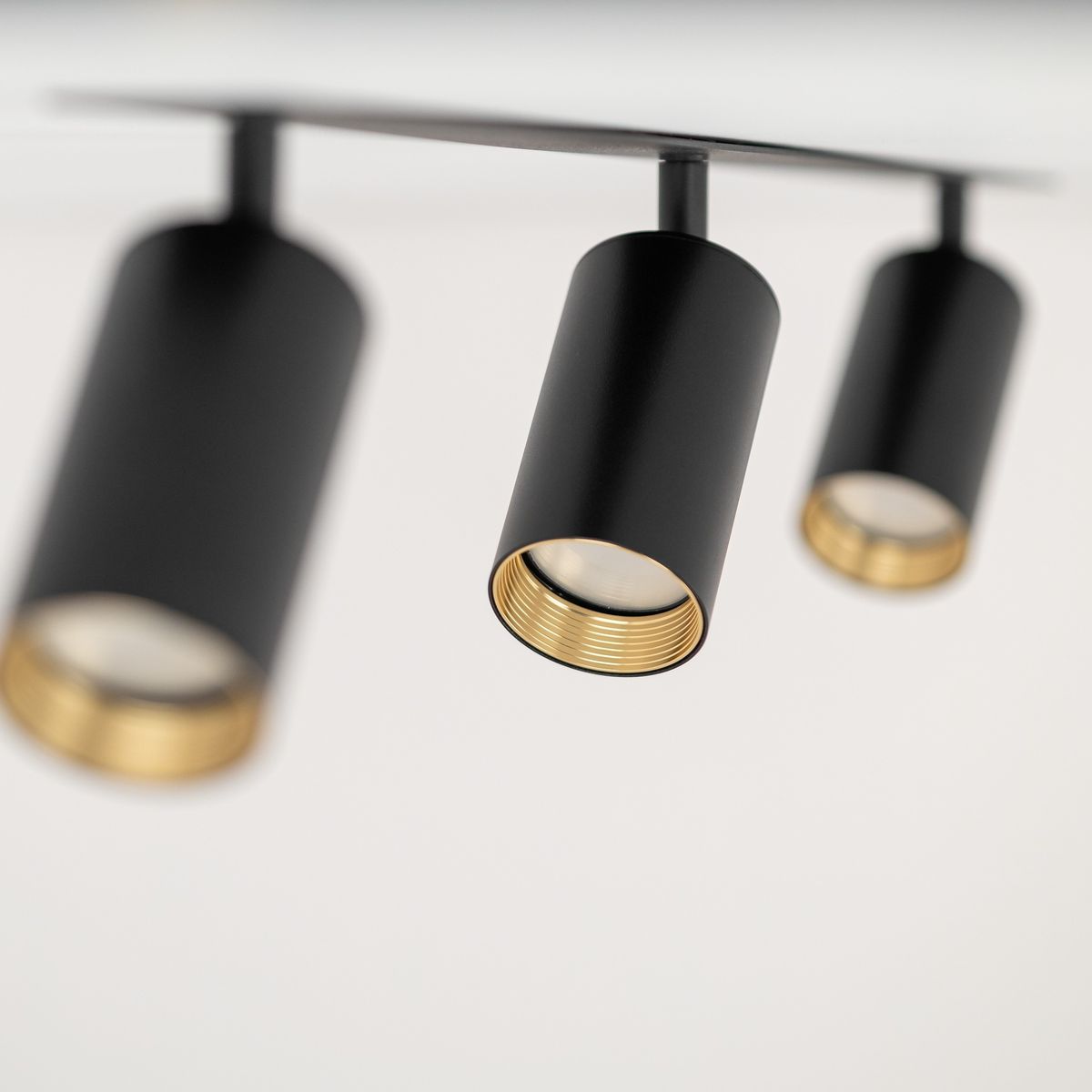 Lampa sufitowa Amis w kolorze czarnym, z trzema spotami na żarówki GU10. Ujęcie z bliska pokazuje cylindryczne klosze z złotymi detalami wewnętrznymi, zamontowane na prostokątnej podstawie.