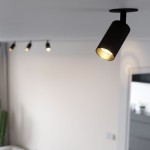 Lampa sufitowa podtynkowa Amis w kolorze czarnym z pojedynczym reflektorem GU10. Ujęcie z góry, pokazujące lampę zamontowaną na suficie, emitującą światło. W tle widoczne inne lampy spot oraz neutralna, jasna ściana.