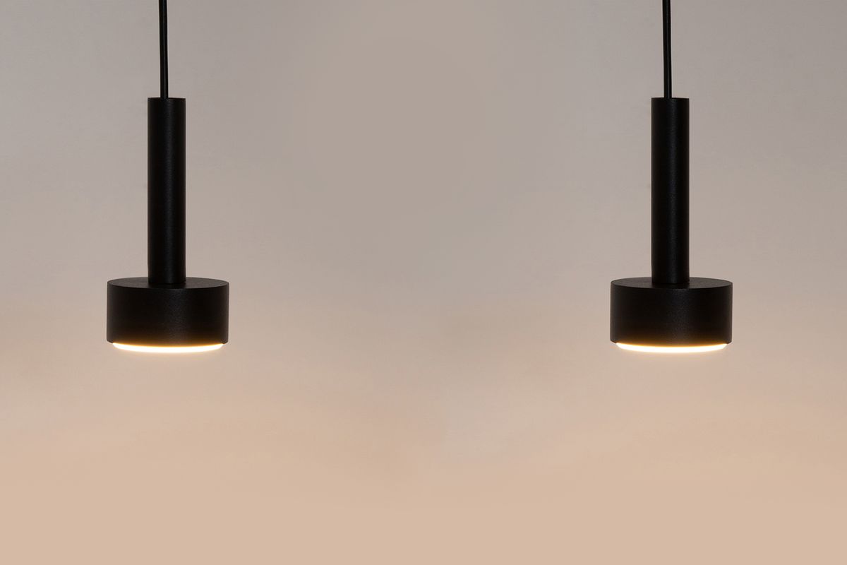 Lampa wisząca Stamp w kolorze czarnym, z dwoma oprawami GX53. Posiada cylindryczny kształt z okrągłą podstawą. Zawieszona na cienkim przewodzie, emitująca delikatne światło. Ujęcie frontalne, na jednolitym tle.