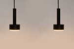 Lampa wisząca Stamp w kolorze czarnym, z dwoma oprawami GX53. Posiada cylindryczny kształt z okrągłą podstawą. Zawieszona na cienkim przewodzie, emitująca delikatne światło. Ujęcie frontalne, na jednolitym tle.