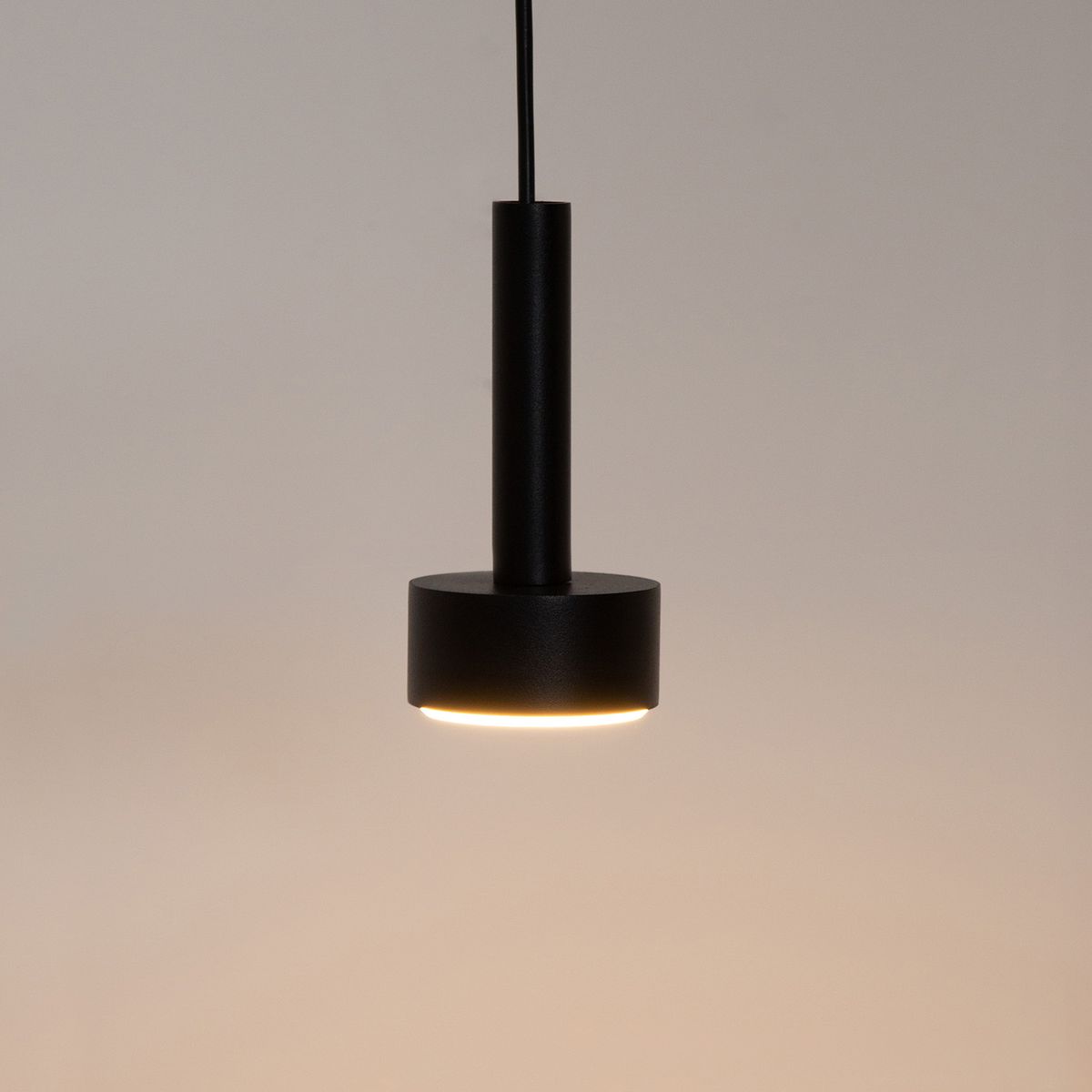 Czarna lampa wisząca typu Stamp z trzema gniazdami GX53, o cylindrycznym kształcie. Produkt zawieszony na cienkim przewodzie, z okrągłą, płaską podstawą emitującą ciepłe światło. Tło proste, neutralne.