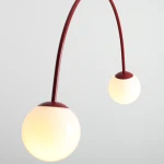 Lampa wisząca Una Red Wine z dwoma kulistymi kloszami w białym kolorze, zawieszona na elastycznym, czerwonym ramieniu. Klosze mają metalowe wykończenie u góry, całość nowoczesna i minimalistyczna, idealna do różnych aranżacji wnętrz.