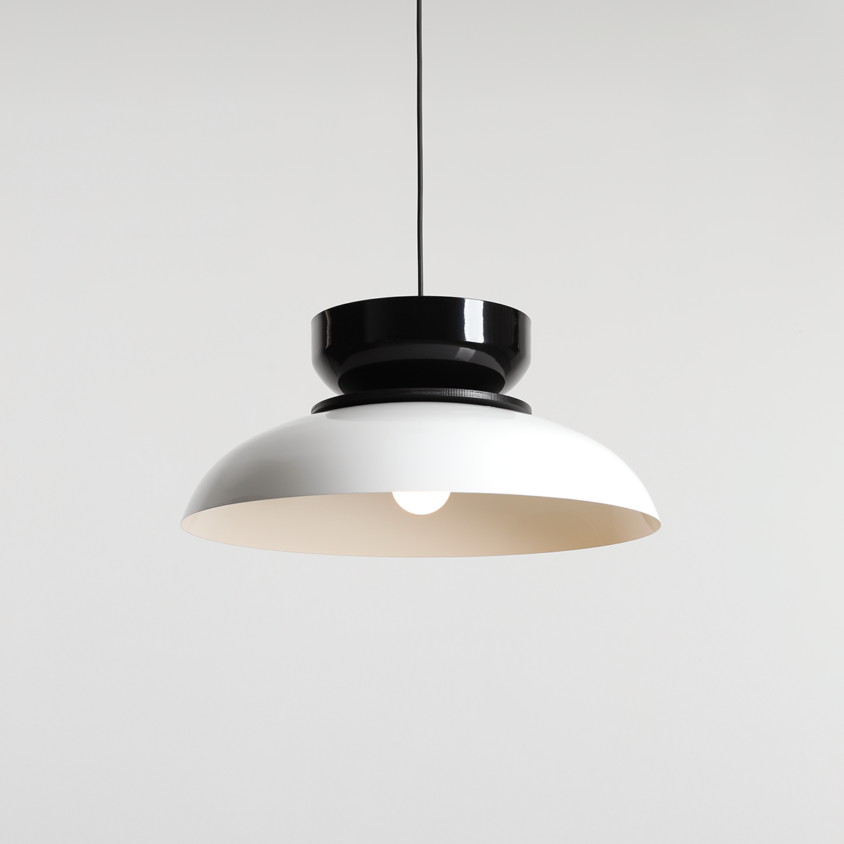 Lampa wisząca Aldo w kolorze białym i czarnym, z metalowym kloszem w kształcie półkuli. Ujęcie frontalne, zawieszona na cienkim przewodzie, z widoczną żarówką E27 w środku. Proste tło podkreśla minimalistyczny design lampy.