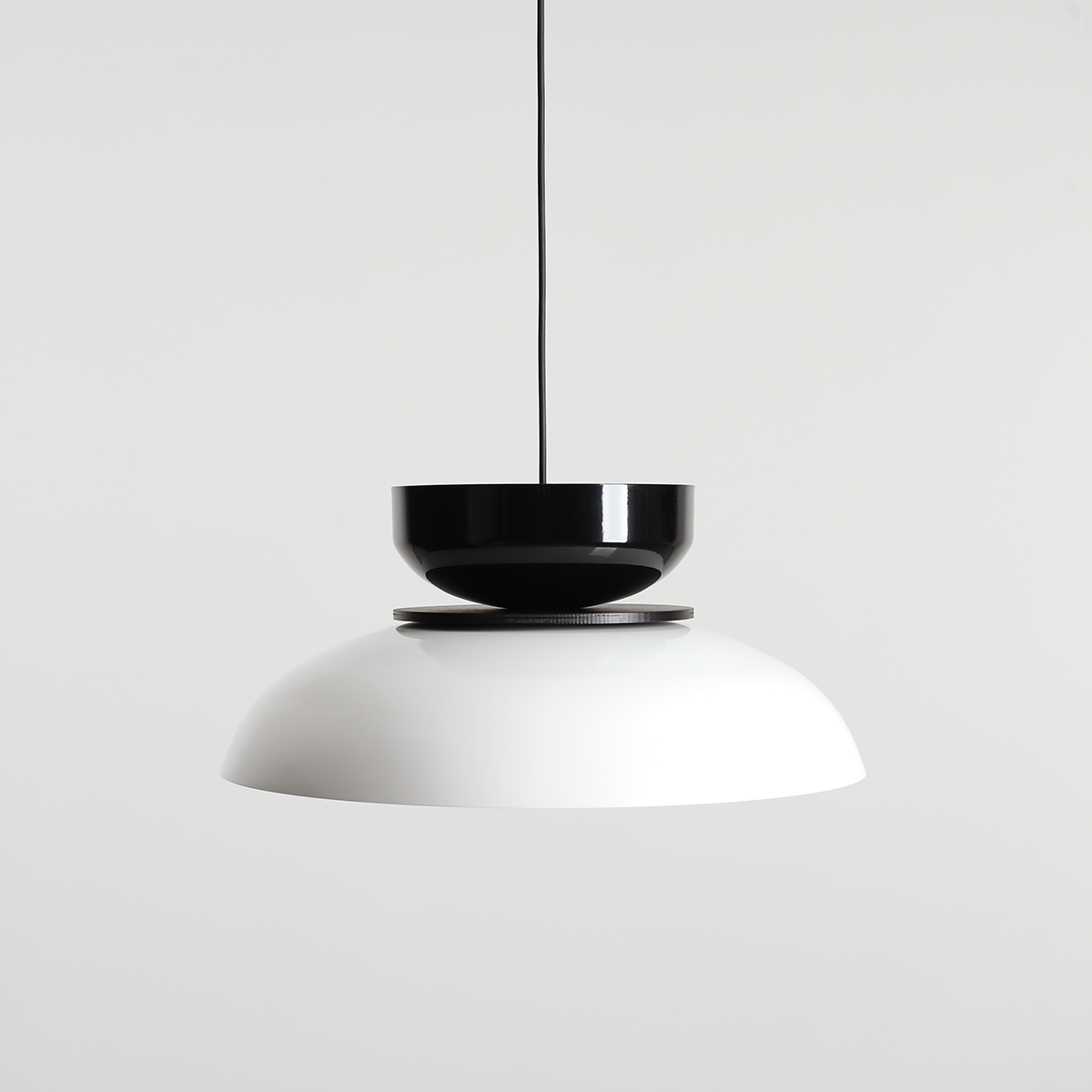 Lampa wisząca Aldo z jedną oprawą E27 w kolorze białym i czarnym. Posiada połyskującą, okrągłą czaszę w białym kolorze oraz czarną, cylindryczną górę. Zawieszona na cienkim, czarnym przewodzie na jasnym tle.