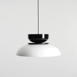 Lampa wisząca Aldo z jedną oprawą E27 w kolorze białym i czarnym. Posiada połyskującą, okrągłą czaszę w białym kolorze oraz czarną, cylindryczną górę. Zawieszona na cienkim, czarnym przewodzie na jasnym tle.